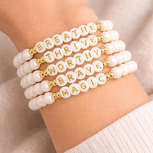 May include: White beaded bracelets with gold lettering spelling out "BREATHE", "THRIVE", "WORTHY", "BRAVE", and "MAGIC". The bracelets are worn on a wrist, with gold accents between the beads.
