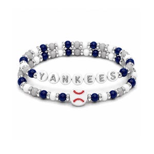 Op de afbeelding: Twee kralenarmbanden met marineblauwe, witte en grijze ronde kralen en zilverkleurige tussenstukjes. Eén armband toont witte letterkralen die YANKEES spellen, terwijl de andere een centrale witte kraal heeft met een rood honkbalstikselpatroon.