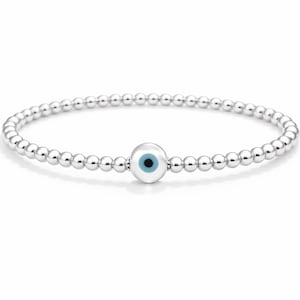 Op de afbeelding: Zilveren kralenarmband met een centrale bedel met een blauw oogontwerp. De armband is gemaakt van kleine, ronde, zilveren kralen en wordt op een witte achtergrond weergegeven.