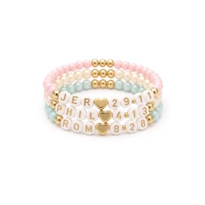 May include: Three beaded bracelets in pastel pink, white, and mint green. Each bracelet features white letter beads spelling out names and numbers, with gold heart and bead accents. A stylish accessory.