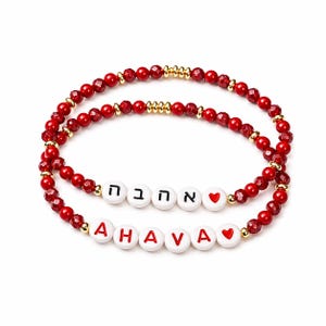 Puede incluir: Dos pulseras de cuentas rojas con detalles dorados. Una pulsera deletrea "AHAVA" en cuentas de letras blancas con corazones rojos. La otra pulsera tiene letras hebreas. Joyería.