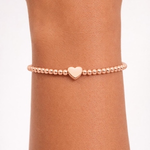 May include: Rose gold beaded bracelet with a heart charm. The bracelet features small, round beads strung together, with a smooth heart-shaped charm centered on the wrist. A simple and elegant piece of jewelry.