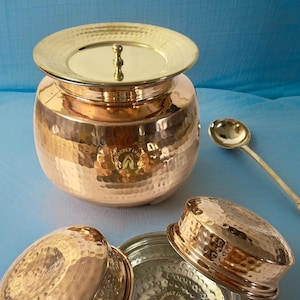 May include: A set of copper kitchenware, including a large pot with a lid, two smaller bowls with lids, and a ladle. The copper is hammered, giving it a textured appearance. The pieces are all polished and shiny.
