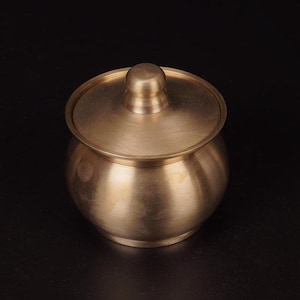 Puede incluir: Un pequeño recipiente redondo de metal de color dorado con tapa. El recipiente tiene una superficie lisa y brillante.