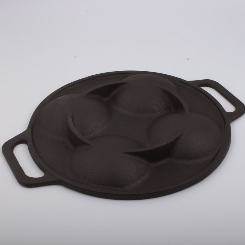Puede incluir: Una sart&eacute;n de hierro fundido negra con cuatro moldes redondos para hacer pasteles individuales u otros productos horneados. La sart&eacute;n tiene dos asas.