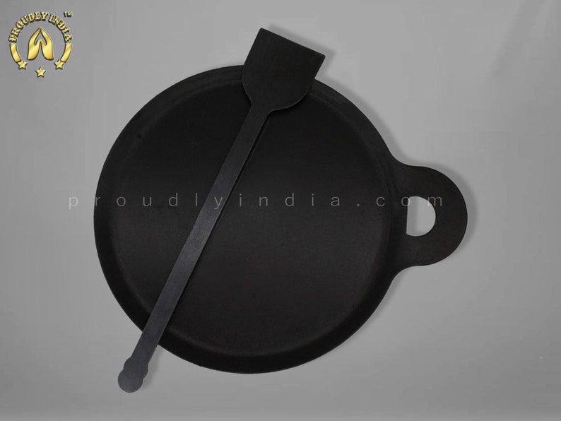 K&ouml;nnte beinhalten: Eine schwarze Gusseisen-Dosa-Pfanne mit einem Griff und einem passenden schwarzen Spatel. Die Pfanne ist rund und hat einen leicht erh&ouml;hten Rand. Der Spatel ist lang und d&uuml;nn mit einem abgerundeten Ende.