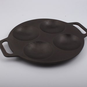 Puede incluir: Una sart&eacute;n de hierro fundido negra con cuatro hendiduras redondas para cocinar porciones individuales de comida. La sart&eacute;n tiene dos asas.