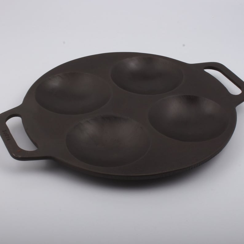 Puede incluir: Una sart&eacute;n de hierro fundido negra con cuatro hendiduras redondas para cocinar porciones individuales de comida. La sart&eacute;n tiene dos asas.