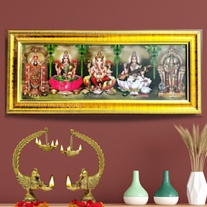 Puede incluir: Una impresi&oacute;n enmarcada de cinco deidades hind&uacute;es, incluyendo Lakshmi, Ganesha, Saraswati y otras dos, en un marco dorado. La impresi&oacute;n est&aacute; en una pared con dos l&aacute;mparas de aceite de lat&oacute;n en forma de pavo real en primer plano. El texto "proudlyindia.com" es visible en la parte inferior de la impresi&oacute;n.