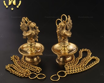 May include: Two gold-colored hanging oil lamps with peacock designs and chains. The lamps are on a black background.
