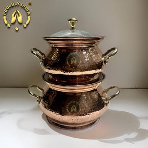 Puede incluir: Un plato de servir de cobre de dos niveles con textura martillada y detalles dorados. El plato tiene dos asas a cada lado y una tapa de vidrio.