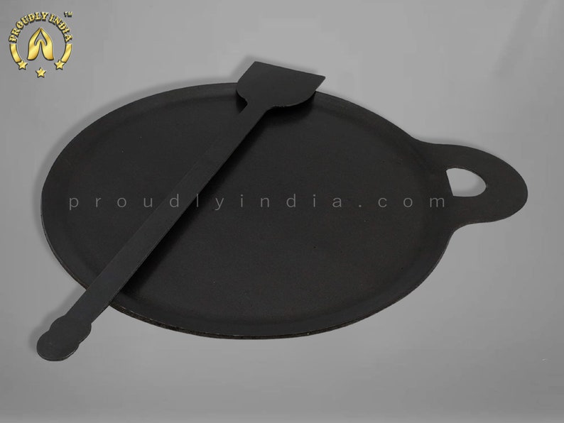 K&ouml;nnte beinhalten: Eine schwarze Gusseisen-Dosa-Pfanne mit einem Griff und einem passenden schwarzen Spatel. Die Pfanne ist rund und hat einen leicht erh&ouml;hten Rand. Der Spatel ist lang und d&uuml;nn mit einem abgerundeten Ende.