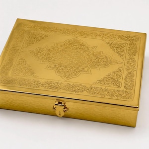 Puede incluir: Una caja rectangular de metal color dorado con tapa abatible. La superficie superior presenta un intrincado patrón grabado con un diseño central trenzado y bordes florales. Los lados tienen una textura martillada y un cierre central.