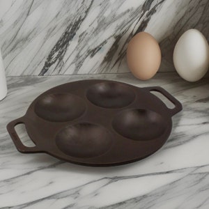 Puede incluir: Una sart&eacute;n de hierro fundido negra con cuatro hendiduras redondas para cocinar huevos. La sart&eacute;n tiene un solo mango en un lado y dos mangos m&aacute;s peque&ntilde;os en el otro.