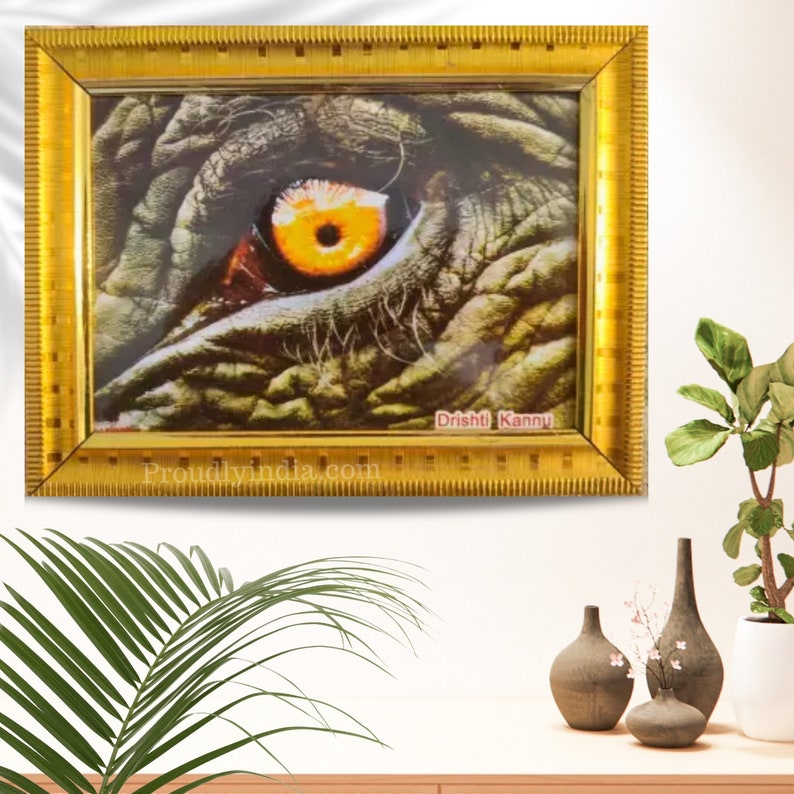Puede incluir: Una impresi&oacute;n enmarcada de un primer plano de un ojo de elefante. El ojo es amarillo y marr&oacute;n con una pupila negra. La impresi&oacute;n est&aacute; enmarcada en un marco dorado con el texto "Dristhi Kannu" y "Proudlyindia.com" en la parte inferior.