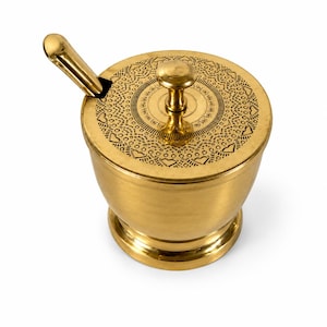 Puede incluir: Un recipiente de metal de color dorado pulido con una tapa a juego y una cuchara pequeña. La tapa presenta un pomo central y un intrincado patrón grabado de pequeños corazones y formas geométricas. El recipiente tiene un cuerpo redondeado y liso.