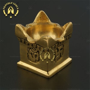 以下が含まれることがあります： 正方形のベースとボウル型のトップを備えた、金色の真鍮製オイルランプ。 ランプにはエンボス加工されたシンボルと「PROUDLY INDIA」の文字が施されています。 暗い表面に置かれています。