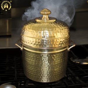Pode incluir: Uma panela de metal martelada dourada com uma tampa e uma pega no topo. O vapor está a subir da panela, que está sobre um fogão preto.
