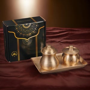 Pode incluir: Um conjunto de dois recipientes de latão com tampas e uma bandeja a condizer, apresentado com uma caixa preta com padrões decorativos dourados. Os recipientes têm uma forma clássica.