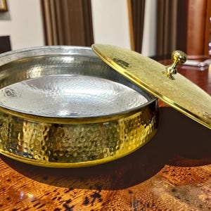 May include: A hammered metal serving bowl with a gold-coloured exterior and a silver-coloured interior. The bowl has a matching lid with a gold knob. The lid is partially open, revealing the inside of the bowl. The bowl is on a wooden surface.