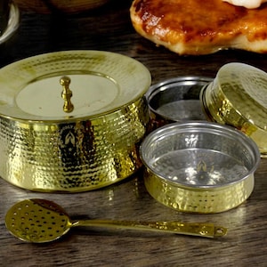 May include: A set of four gold-toned hammered metal bowls with lids. The bowls are of varying sizes and are arranged on a wooden surface. A small gold-toned slotted spoon is also included.