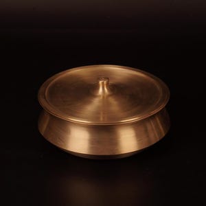 Puede incluir: Una olla redonda de latón con tapa. La olla tiene una superficie lisa y brillante y un borde ligeramente acampanado. La tapa tiene un pequeño pomo en la parte superior.
