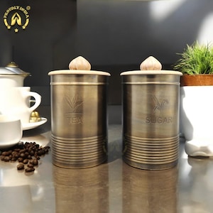 Puede incluir: Dos botes de metal dorado antiguo con tapas de madera. El bote de la izquierda está etiquetado con "TEA" y el bote de la derecha está etiquetado con "SUGAR".