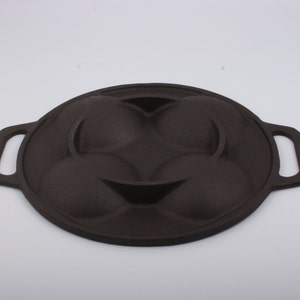 Puede incluir: Una sart&eacute;n de hierro fundido negra con cuatro moldes redondos para hacer pasteles individuales u otras golosinas. La sart&eacute;n tiene dos asas a los lados.