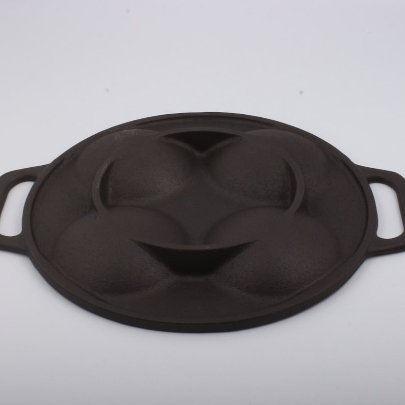 Puede incluir: Una sart&eacute;n de hierro fundido negra con cuatro moldes redondos para hacer pasteles individuales u otras golosinas. La sart&eacute;n tiene dos asas a los lados.