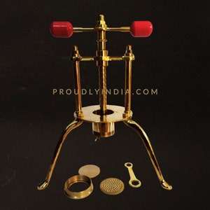 Può includere: Una macchina per la pasta in metallo dorato con manici rossi e tre gambe. Include una base rotonda, un disco forato e un anello.