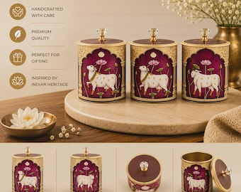 May include: Three cylindrical metal storage canisters with lids, featuring a deep magenta background, gold-toned decorative arches, and a white cow illustration surrounded by lotus flowers. Each lid is topped with a clear crystal-like knob.