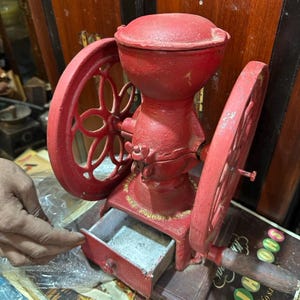 May include: A vintage, red cast-iron coffee grinder with two large wheels and a drawer. The grinder has a hopper on top and a wooden handle. The drawer is open, revealing the interior. The grinder is sitting on a stack of vintage magazines.