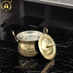 May include: A gold-colored metal pot with a lid. The pot has a round body and two handles. The lid has a knob on top. The pot is sitting on a black marble surface.