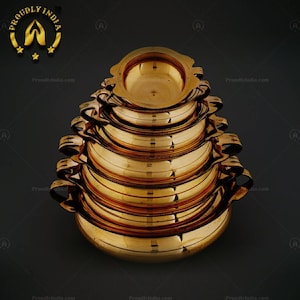 May include: A set of six stacked brass bowls with rounded edges and handles. The bowls are arranged in a pyramid shape, with the largest bowl at the bottom and the smallest bowl at the top.