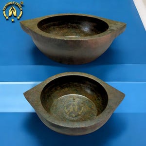 May include: Two antique-style, dark bronze-coloured bowls with a unique, eye-shaped design. The bowls have a textured surface and a wide, open interior. The bowls are displayed on a blue surface, with a logo in the upper left corner.