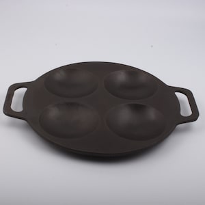 Puede incluir: Una sart&eacute;n de hierro fundido negra con cuatro hendiduras redondas para cocinar porciones individuales de comida. La sart&eacute;n tiene dos asas.