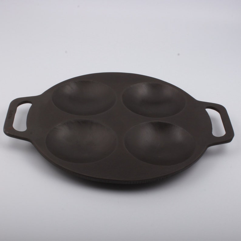 Puede incluir: Una sart&eacute;n de hierro fundido negra con cuatro hendiduras redondas para cocinar porciones individuales de comida. La sart&eacute;n tiene dos asas.