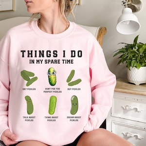 May include: Pink sweatshirt with the text "THINGS I DO IN MY SPARE TIME" and a series of cartoon pickles with different expressions and activities, such as "EAT PICKLES", "HUNT FOR THE PERFECT PICKLES", "BUY PICKLES", "TALK ABOUT PICKLES", "THINK ABOUT PICKLES", and "DREAM ABOUT PICKLES".