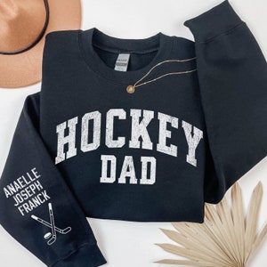 May include: Black sweatshirt with the text "HOCKEY DAD" in white on the front. The left sleeve has the text "ANAELLE JOSEPH FRANCK" and a hockey stick graphic in white.