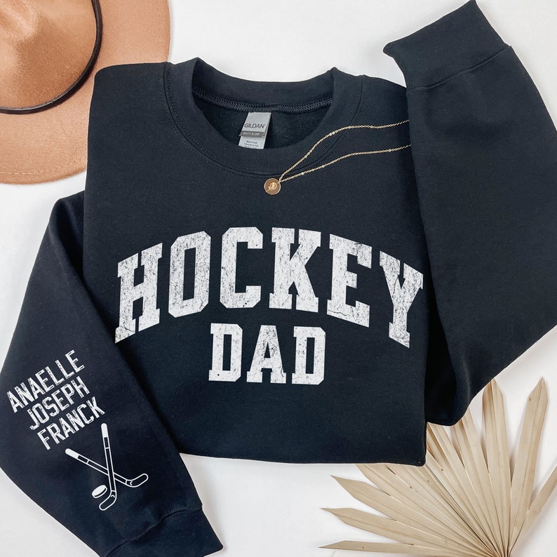 May include: Black sweatshirt with the text "HOCKEY DAD" in white on the front. The left sleeve has the text "ANAELLE JOSEPH FRANCK" and a hockey stick graphic in white.