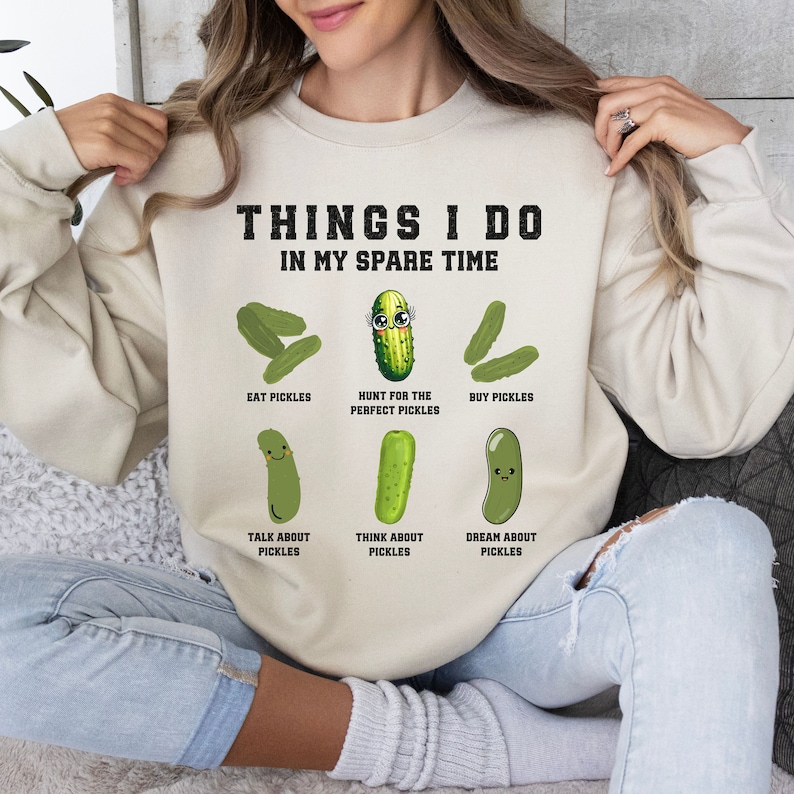 May include: A beige sweatshirt with the text "THINGS I DO IN MY SPARE TIME" and a series of illustrations of pickles with different expressions and activities, such as "EAT PICKLES", "HUNT FOR THE PERFECT PICKLES", "BUY PICKLES", "TALK ABOUT PICKLES", "THINK ABOUT PICKLES", and "DREAM ABOUT PICKLES".