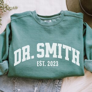 May include: A teal green sweatshirt with white text that reads "DR. SMITH EST. 2023".