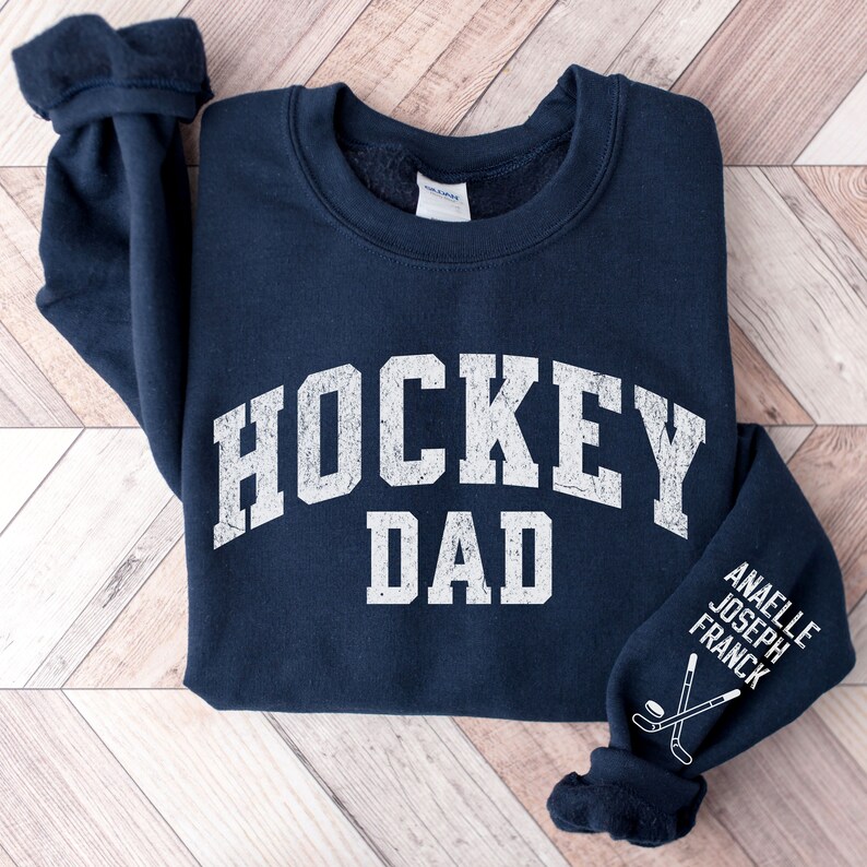 May include: Navy blue sweatshirt with the text "HOCKEY DAD" in white on the front. The left sleeve has the text "ANAELLE JOSEPH FRANCK" and a hockey stick and puck graphic in white.