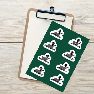 May include: Nine white stickers with a black and gray illustration of a person wearing a black mask and using a laptop. The text "DATA NINJA" is written in black below the illustration on each sticker. The stickers are arranged in a grid pattern on a green background.