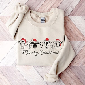May include: A beige sweatshirt with a Christmas-themed design featuring four cows wearing Santa hats and the text "Moo-ry Christmas".