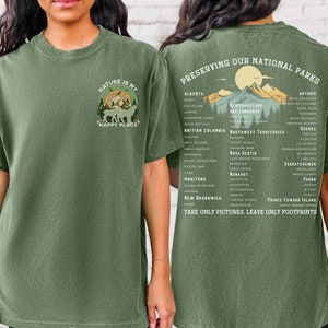 May include: Green t-shirt with a nature-themed design on the front and a list of Canadian national parks on the back. The front design features a forest scene with the text "Nature is my happy place". The back design features a mountain range with the text "Preserving our National Parks" and a list of parks by province.