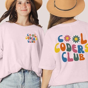 May include: Pink t-shirt with a retro-style graphic that says "COOL CODERS CLUB" in colourful letters with flowers. The graphic is on the back of the shirt.