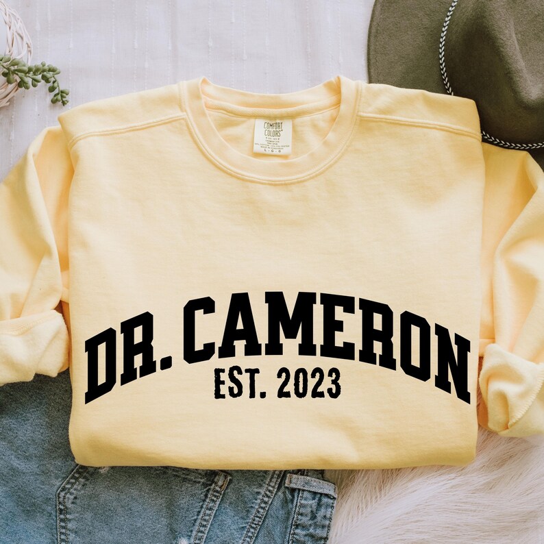 May include: A yellow sweatshirt with black text that reads "Dr. Cameron Est. 2023".