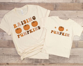 May include: Two cream coloured t-shirts with a pumpkin theme. The larger shirt says "Raising Pumpkins" with three pumpkins in a row. The smaller shirt says "Pumpkin" with one pumpkin and two stars.