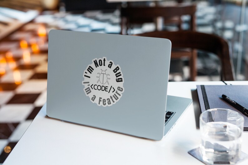 Pode incluir: Um laptop prateado com um adesivo que diz "Eu n&atilde;o sou um bug, eu sou um recurso <CODE/>" com um bug de desenho animado dentro do c&oacute;digo.